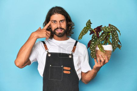 Téléchargez les photos : Jardinier aux cheveux longs avec barbe tenant une plante montrant un geste de déception avec index. - en image libre de droit
