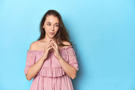 Téléchargez les photos : Jeune femme vêtue d'une robe rose sur fond de studio bleu maquillant le plan à l'esprit, mettant en place une idée. - en image libre de droit