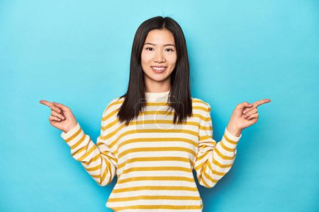 Photo for Asian woman in striped yellow sweater, pointing to different copy spaces, choosing one of them, showing with finger. - Royalty Free Image