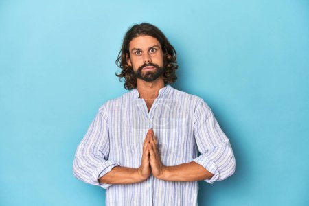 Photo for Man with beard in blue striped shirt, blue studio praying, showing devotion, religious person looking for divine inspiration. - Royalty Free Image