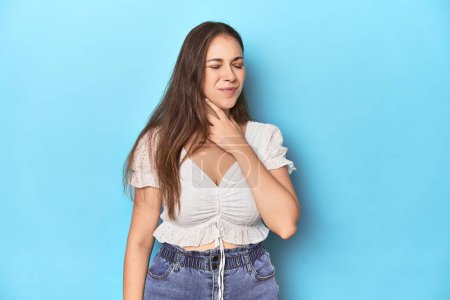 Téléchargez les photos : Jeune femme élégante en chemisier blanc sur fond de studio bleu souffre de douleur à la gorge due à un virus ou une infection. - en image libre de droit