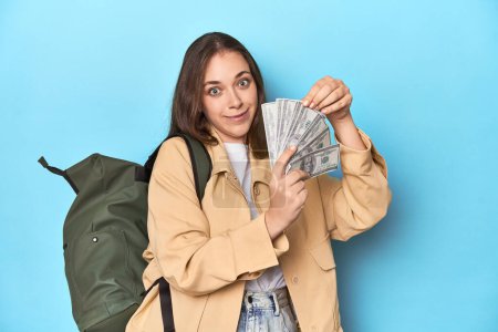 Téléchargez les photos : Femme voyageuse avec sac à dos tenant des billets de dollar sur bleu. - en image libre de droit