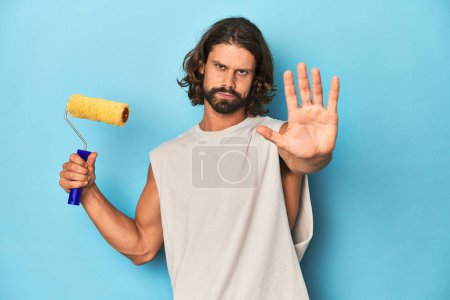 Téléchargez les photos : Peinture homme barbu avec un rouleau jaune debout avec la main tendue montrant le signe d'arrêt, vous empêchant. - en image libre de droit