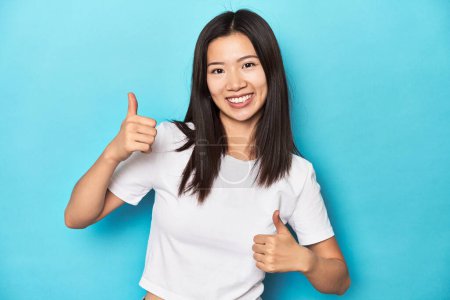 Foto de Mujer asiática joven en camiseta blanca, tiro al estudio, levantando ambos pulgares hacia arriba, sonriendo y confiado. - Imagen libre de derechos