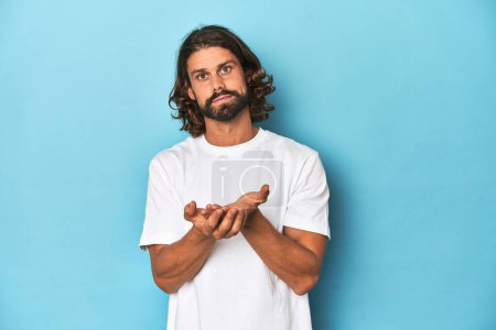 Photo for Bearded man in a white shirt, blue backdrop holding something with palms, offering to camera. - Royalty Free Image