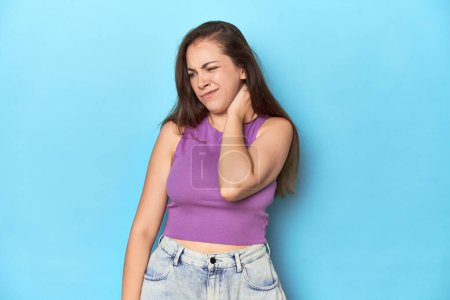 Photo for Fashionable young woman in a purple top on blue background having a neck pain due to stress, massaging and touching it with hand. - Royalty Free Image