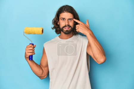 Photo for Bearded man painting with a yellow roller showing a disappointment gesture with forefinger. - Royalty Free Image