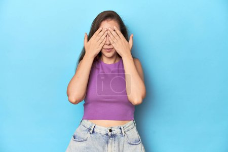 Photo for Fashionable young woman in a purple top on blue background afraid covering eyes with hands. - Royalty Free Image