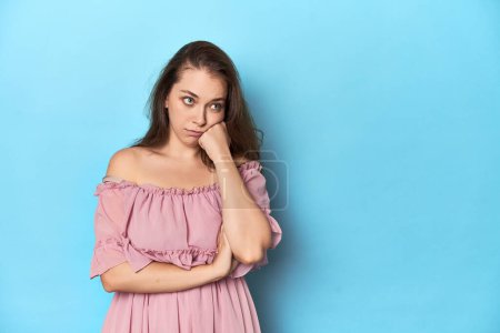Téléchargez les photos : Jeune femme portant une robe rose sur fond de studio bleu qui se sent triste et onéreux, en regardant l'espace de copie. - en image libre de droit