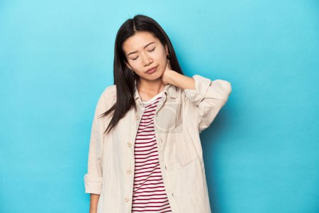 Foto de Mujer asiática con camisa en capas y camiseta a rayas, sufriendo dolor de cuello debido a un estilo de vida sedentario. - Imagen libre de derechos
