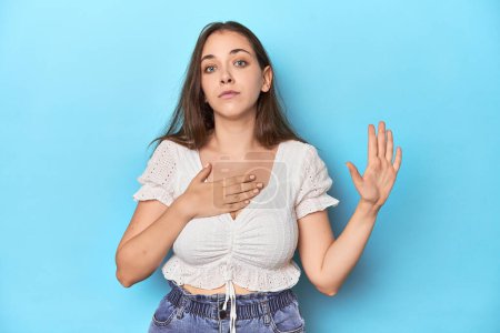 Photo for Stylish young woman in white blouse on a blue studio backdrop taking an oath, putting hand on chest. - Royalty Free Image