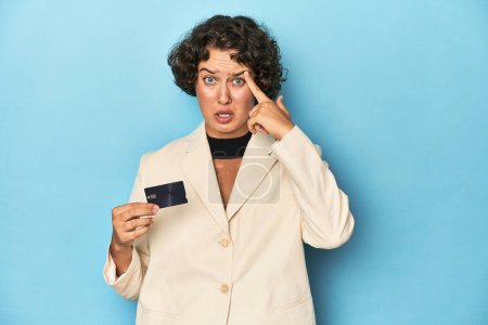 Foto de Mujer joven con tarjeta de crédito, elegante chaqueta blanca que muestra un gesto de decepción con el dedo índice. - Imagen libre de derechos