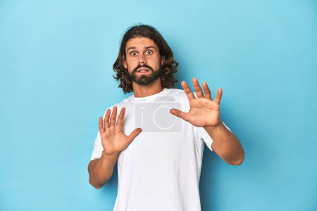 Photo for Bearded man in a white shirt, blue backdrop being shocked due to an imminent danger - Royalty Free Image