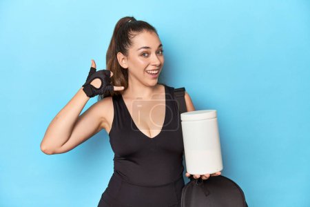 Foto de Mujer joven sosteniendo la botella de proteína en un entorno deportivo que muestra un gesto de llamada de teléfono móvil con los dedos. - Imagen libre de derechos