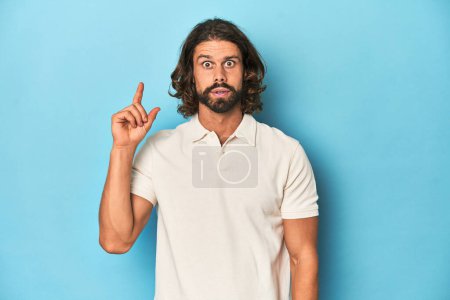 Téléchargez les photos : Homme aux cheveux longs dans un polo blanc, studio bleu ayant une idée, concept d'inspiration. - en image libre de droit