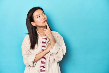 Téléchargez les photos : Femme asiatique en chemise à couches et t-shirt rayé, regardant latéralement avec une expression douteuse et sceptique. - en image libre de droit