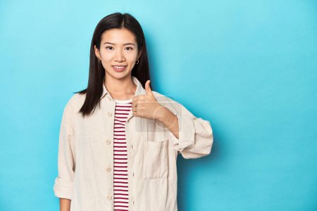 Foto de Mujer asiática con camisa en capas y camiseta a rayas, sonriendo y levantando el pulgar hacia arriba - Imagen libre de derechos