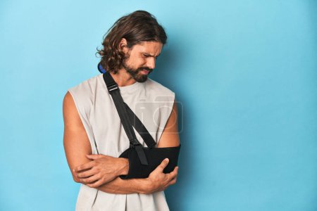 Photo for Man with a beard, long hair and arm in sling looking pained in blue studio. - Royalty Free Image