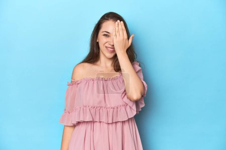 Téléchargez les photos : Jeune femme portant une robe rose sur fond de studio bleu s'amusant à couvrir la moitié du visage avec de la paume. - en image libre de droit