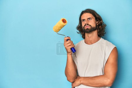 Photo for Bearded man holding a paint roller in a blue studio setting. - Royalty Free Image