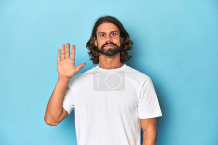 Foto de Hombre barbudo con camisa blanca, fondo azul sonriente alegre mostrando el número cinco con los dedos. - Imagen libre de derechos