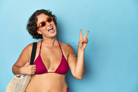 Photo for Young woman in bikini with beach bag joyful and carefree showing a peace symbol with fingers. - Royalty Free Image