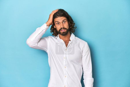 Photo for Bearded man in a white shirt, blue backdrop being shocked, she has remembered important meeting. - Royalty Free Image