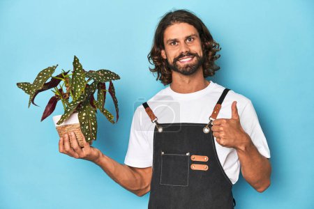 Foto de Jardinero de pelo largo con barba sosteniendo una planta sonriendo y levantando el pulgar hacia arriba - Imagen libre de derechos