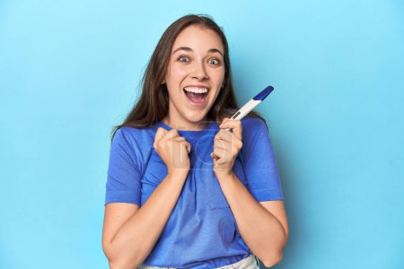 Photo for Excited woman holding a positive pregnancy test on blue studio. - Royalty Free Image