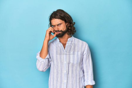Foto de Hombre con barba en camisa de rayas azules, estudio azul con los dedos en los labios manteniendo un secreto. - Imagen libre de derechos