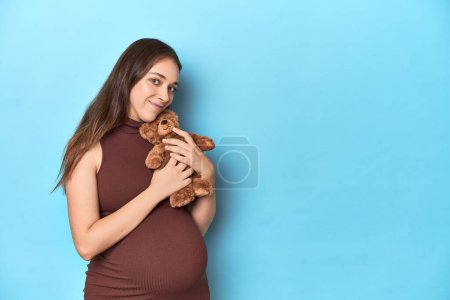 Foto de Mujer embarazada sosteniendo un juguete de bebé de felpa en un estudio azul. - Imagen libre de derechos