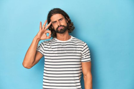 Photo for Bearded man in a striped shirt, blue backdrop cheerful and confident showing ok gesture. - Royalty Free Image
