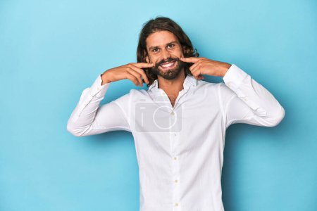 Foto de Hombre barbudo con una camisa blanca, fondo azul sonríe, señalando con los dedos a la boca. - Imagen libre de derechos