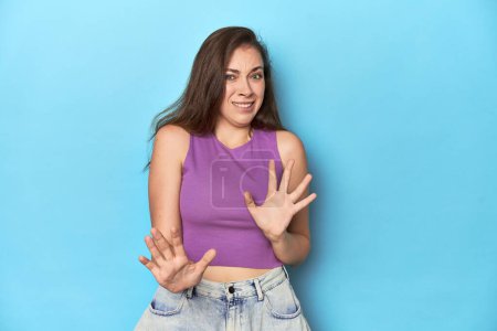 Photo for Fashionable young woman in a purple top on blue background rejecting someone showing a gesture of disgust. - Royalty Free Image