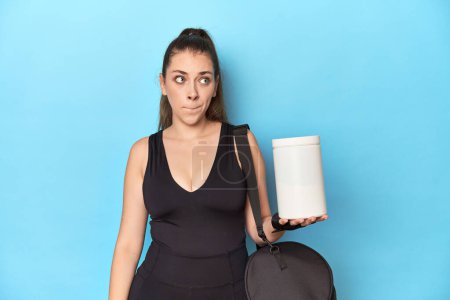 Photo for Young woman holding protein bottle in sporty setting confused, feels doubtful and unsure. - Royalty Free Image