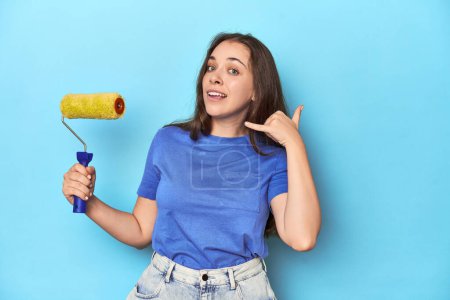 Photo for Young woman with yellow paint roller on a blue background showing a mobile phone call gesture with fingers. - Royalty Free Image