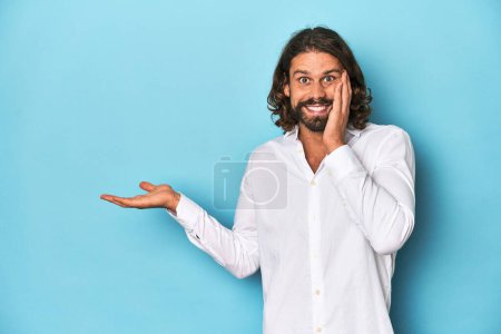 Photo for Bearded man in a white shirt, blue backdrop holds copy space on a palm, keep hand over cheek. Amazed and delighted. - Royalty Free Image