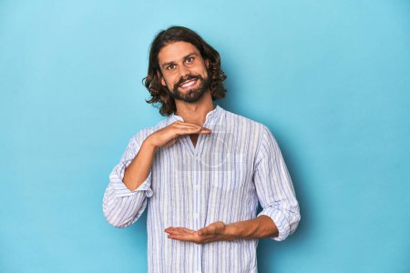 Photo for Man with beard in blue striped shirt, blue studio holding something with both hands, product presentation. - Royalty Free Image