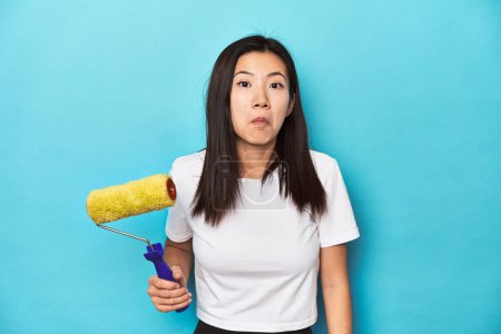 Photo for Young Asian woman with paint roller, DIY concept, shrugs shoulders and open eyes confused. - Royalty Free Image