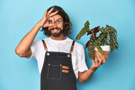 Foto de Jardinero de pelo largo con barba sosteniendo una planta excitada manteniendo un buen gesto a los ojos. - Imagen libre de derechos