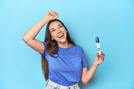 Foto de Mujer emocionada sosteniendo una prueba de embarazo positiva en estudio azul. - Imagen libre de derechos