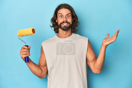 Photo for Bearded man painting with a yellow roller surprised and shocked. - Royalty Free Image