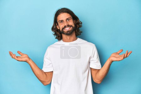 Photo for Bearded man in a white shirt, blue backdrop makes scale with arms, feels happy and confident. - Royalty Free Image