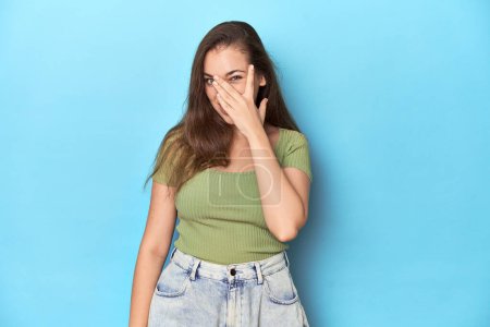 Photo for Young Caucasian woman in a green top on a blue backdrop blink at the camera through fingers, embarrassed covering face. - Royalty Free Image