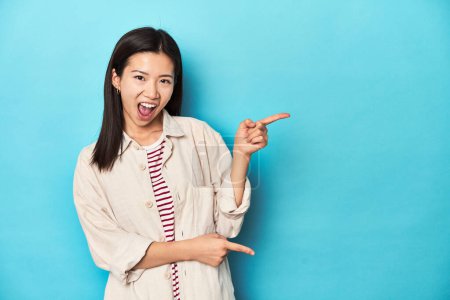Foto de Mujer asiática con camisa en capas y camiseta a rayas, señalando con los dedos delanteros a un espacio de copia, expresando emoción y deseo. - Imagen libre de derechos