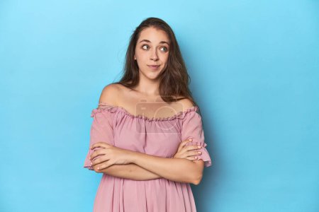 Téléchargez les photos : Jeune femme portant une robe rose sur fond de studio bleu rêvant d'atteindre des objectifs et des buts - en image libre de droit