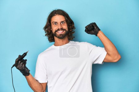 Photo for Bearded tattooist with tattoo machine at work raising fist after a victory, winner concept. - Royalty Free Image