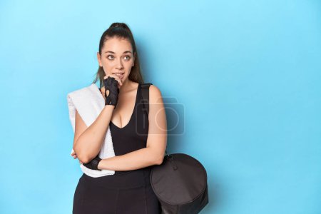 Photo for Young woman ready for gym with essentials on blue backdrop relaxed thinking about something looking at a copy space. - Royalty Free Image