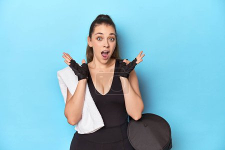 Photo for Young woman ready for gym with essentials on blue backdrop surprised and shocked. - Royalty Free Image
