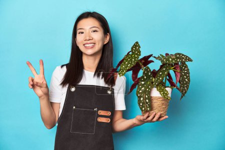 Foto de Joven jardinero asiático sosteniendo la planta, fondo del estudio, mostrando el número dos con los dedos. - Imagen libre de derechos